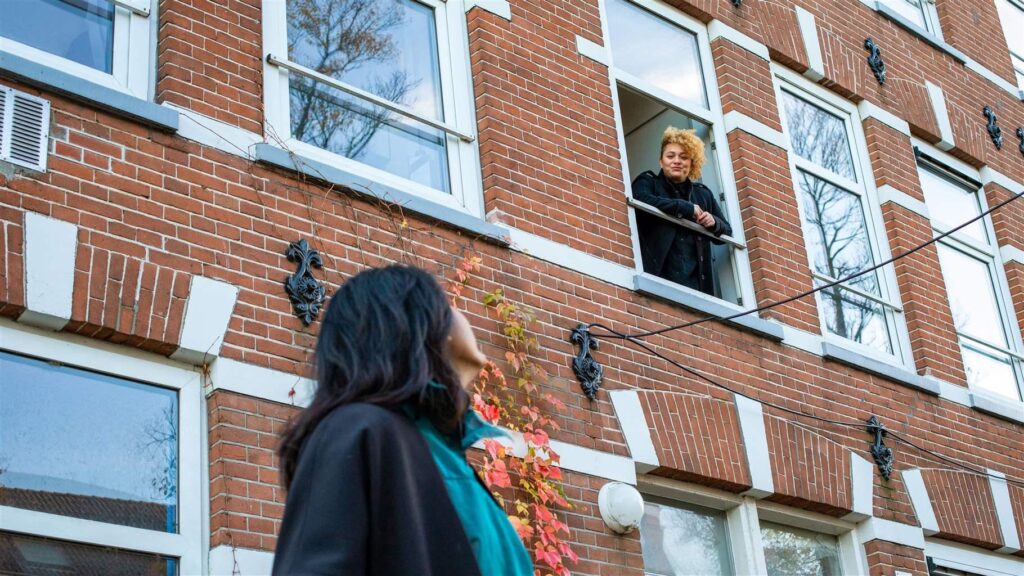 Young person talking through the second-floor window of a brick apartment building with a friend standing outside.